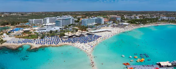 Kıbrıs Rum Kesimi 'nin Ayia Napa kentindeki Nissi Sahili' nin havadan görünüşü, turkuvaz suları, yüzücüler, mavi şemsiyeler, lüks oteller ve arka planda yemyeşil alanlar..