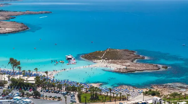 Kıbrıs Rum Kesimi 'nin Ayia Napa kentindeki Nissi Plajı' nın hava manzarasında turkuaz sular, kumlu kıyılar, kayalık bir ada, güneşli yataklar, tatil beldeleri ve su faaliyetleri yer alıyor..