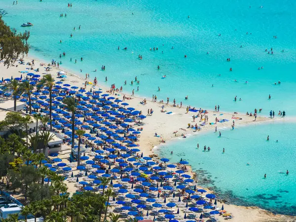 Kıbrıs Rum Kesimi 'nin Ayia Napa kentindeki Nissi Sahili' nin hava manzarasında turkuaz sular, yüzücüler, mavi şemsiyeler, güneş panzeleri, palmiye ağaçları ve yemyeşil alanlar görülüyor..