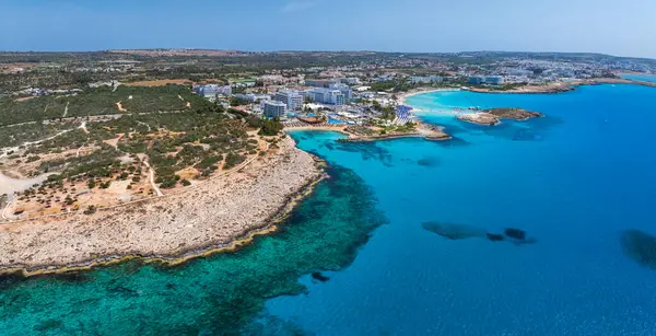 Kıbrıs Rum Kesimi 'nin Ayia Napa kentinin turkuaz suları, kumlu Nissi Sahili, lüks tatil beldeleri, engebeli kıyı şeridi ve yeşillikli kentsel yerleşim yerlerini gösteren hava görüntüleri..