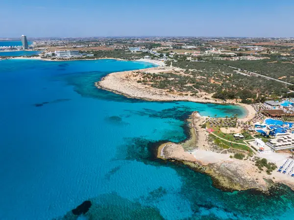 Kıbrıs Rum Kesimi 'nin Ayia Napa kentinin turkuaz sular, kumlu plajlar, lüks tatil beldeleri, havuzlar, kayalık çıkıntılar ve yüksek modern bir bina gösteren hava görüntüsü..