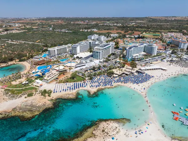Ayia Napa, Kıbrıs Rum Kesimi, Nissi ve Landa Sahillerini, turkuaz suları, lüks tatil beldeleri, güneş pansiyonları, yüzücüler ve çevresindeki manzaraları gösteren hava manzarası.