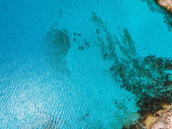 Kıbrıs Rum Kesimi 'nin Ayia Napa kenti yakınlarındaki turkuaz sularda, sualtı özelliklerini gösteren koyu renkli yamalar görülüyor. Sağ üst köşede engebeli, kayalık bir kıyı şeridi görünüyor..