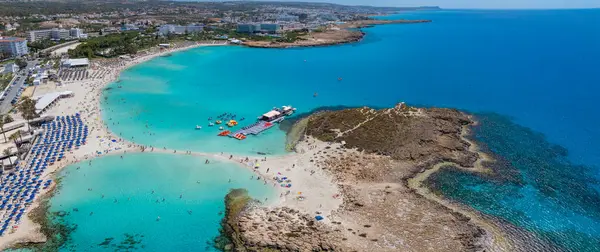 Kıbrıs Rum Kesimi 'nin Ayia Napa kentindeki Nissi Plajı' nın hava manzarasında turkuaz sular, altın kumlar, güneş pansiyonları, su sporları, lüks tatil beldeleri ve kayalık çıkıntılar görülüyor..