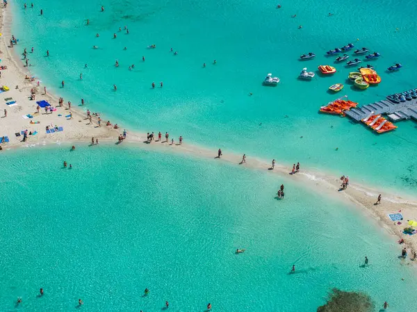 Kıbrıs 'ın Ayia Napa kentindeki turkuaz sular ve kumlu Nissi plajı, bir kum kayığı, yüzücüler, bir rıhtım ve berrak sığ sularda renkli yüzgeçli kayıklar..