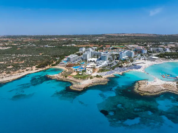 Ayia Napa, Kıbrıs Rum Kesimi, Nissi Sahili, turkuaz sular, lüks tatil beldeleri, güneşlikler, şemsiyeler, yemyeşil alanlar ve kentsel manzaraların yer aldığı hava manzarası.
