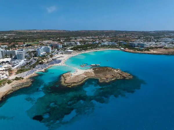 Kıbrıs Rum Kesimi 'nin Ayia Napa kentindeki Nissi Sahili' nin hava manzarasında turkuaz sular, altın kumlar, güneş pansiyonları, yakınlardaki tatil beldeleri ve yüzen insanlar yer alıyor..