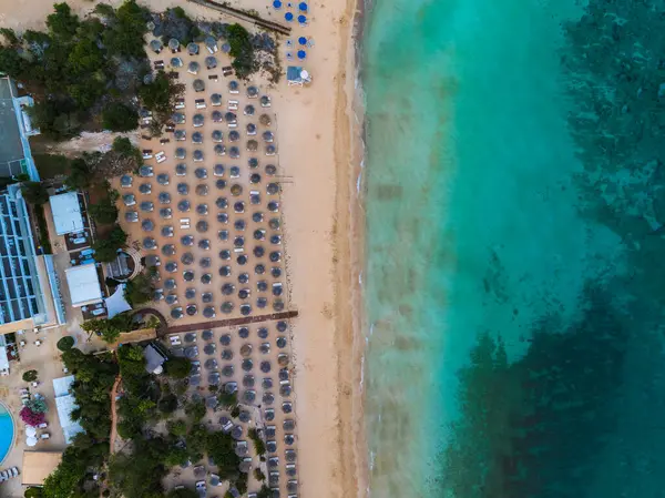 Kıbrıs 'ın Ayia Napa kentinin kumlu bir plajı, turkuaz suları, güneş pansiyonları, şemsiyeler, yakınlardaki tatil beldeleri ve yemyeşil alanları gösteren hava görüntüleri..