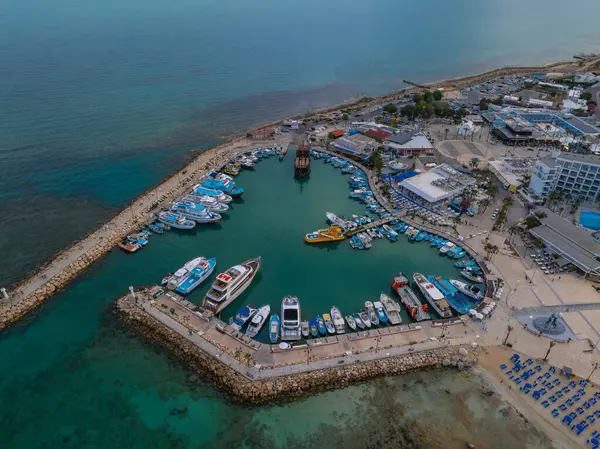 Kıbrıs Rum Kesimi 'nin Ayia Napa kentindeki tekneler, yatlar, modern binalar, kumlu kumsallar, mavi güneş pansiyonları ve turkuaz Akdeniz sularıyla dolu bir marina manzarası..