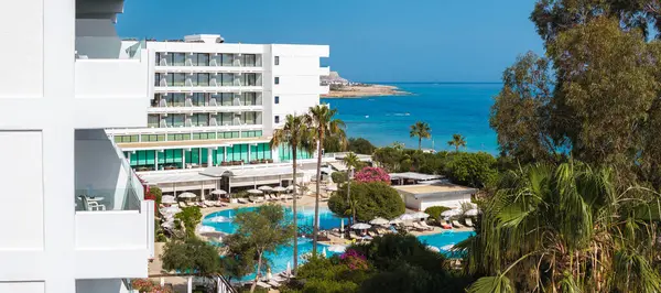 Mavi havuzlu lüks beyaz bir otel, güneş pansiyonları, palmiye ağaçları ve çiçek açan çalılar, turkuaz Akdeniz ve kayalık kıyı şeridi yakınında geçiyor..