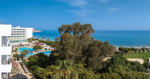 Ayia Napa 'nın beyaz çok katlı oteli, büyük havuzu, güneşli dinlenme yerleri, yemyeşil, kumlu plajı ve turkuaz Akdeniz suları..