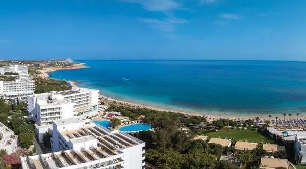 Kıbrıs Rum Kesimi 'nin Ayia Napa kentinin hava manzarasında turkuaz sular, kumlu plajlar, lüks tatil beldeleri, güneşli gökyüzü altında güneşli dinlenme yerleri, şemsiyeler ve yemyeşil alanlar görülüyor..