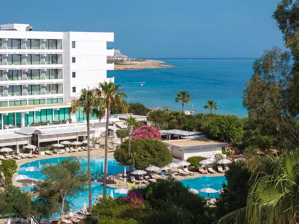 Kıbrıs Rum Kesimi 'nin Ayia Napa kentindeki lüks tatil beldesinde beyaz çok katlı bir otel, canlı bir havuz alanı, yemyeşil ve arka planda Akdeniz yer alıyor..