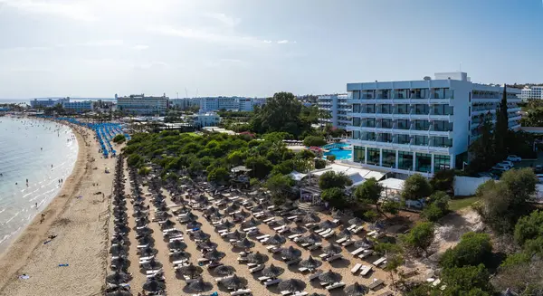 Kıbrıs Rum Kesimi 'nin Ayia Napa kentinin hava manzarası, havuzlu beyaz lüks bir otel, güneş pansiyonlu plaj, turkuaz deniz ve kıyı tatil beldeleri gösteriyor..