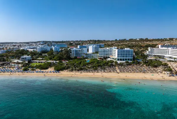 Kıbrıs Rum Kesimi 'nin Ayia Napa kentinin hava manzarasında turkuaz sular, kumlu plajlar, lüks oteller, yemyeşil alanlar ve canlı kıyı boyunca yüzen insanlar görülüyor..