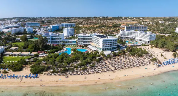 Kıbrıs Rum Kesimi 'nin Ayia Napa kentinin havadan görünüşü, Nissi Sahili' ni, turkuaz suları, güneşlik yatakları, şemsiyeler, lüks tatil beldeleri, havuzlar, bahçeler ve yemyeşil alanları gösteriyor..