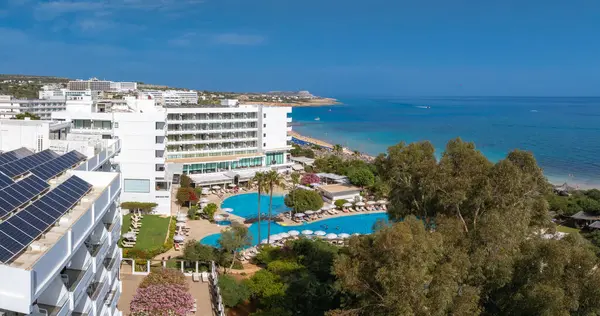 Kıbrıs Rum Kesimi 'nin Ayia Napa kentinin havadan görünüşü, lüks tatil beldeleri, güneşli bir havuz, kumlu plaj, plaja gidenler ve turkuaz Akdeniz sularını gösteriyor..