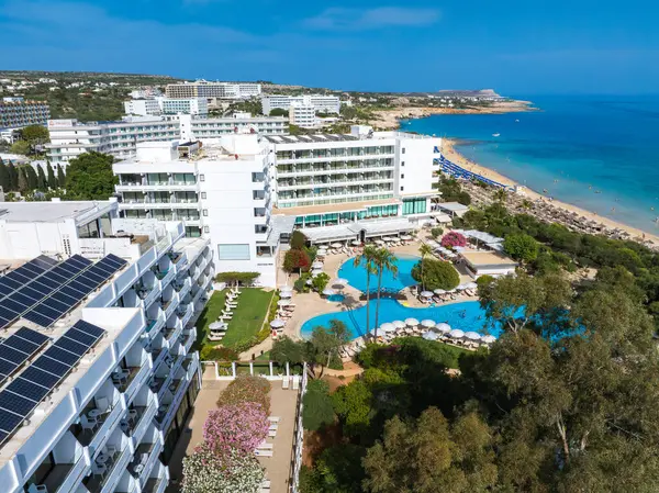 Kıbrıs Rum Kesimi 'nin Ayia Napa kentinin havadan görünüşü, lüks tatil beldeleri, büyük bir havuz, güneş panelleri, Nissi Sahili' nin altın kumları, turkuaz suları ve plaja gidenleri gösteriyor..