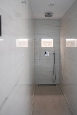 Beyaz mermer duvarları olan gösterişli bir duş alanı, aydınlatılmış oyuklar, bir sağanak duş başlığı, el bilgisayarı, ve zeminde doğrusal bir drenaj..