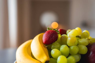 Canlı bir meyve aranjmanı, muzlu, yeşil üzümlü ve odak noktasında kırmızı çilekli. Ilık ışıklandırma ve yumuşak bulanık arkaplan bileşimi artırır.