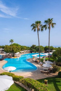 Kıbrıs 'ta bir tatil köyü; kavisli bir havuz, güneşli şezlonglar, şemsiyeler, yüksek palmiye ağaçları, gür yeşillikler ve berrak bir gökyüzünün altındaki mavi deniz..