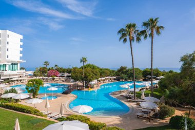 Kıbrıs Rum Kesimi 'nde bir havuz, güneş şezlongları, şemsiyeler, yüksek palmiye ağaçları, yemyeşil yemyeşil, modern beyaz bir otel ve arka planda deniz yer alıyor..