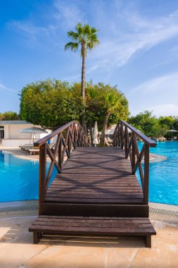 Bir tahta köprü, palmiye ağaçları, tropik bitkiler, beyaz binalar ve şemsiyelerin altındaki şezlonglarla çevrili bir Kıbrıs tatil beldesindeki berrak mavi havuzu geçer..