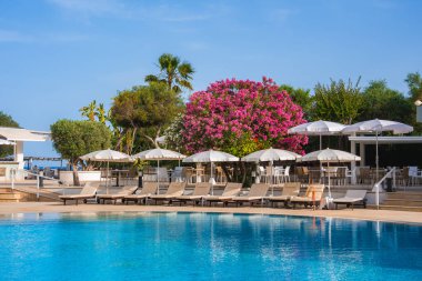 Beyaz şemsiyeli ve şezlonglu berrak mavi bir havuz. Etrafı palmiye ağaçları ve pembe çiçekli ağaçlarla çevrili..