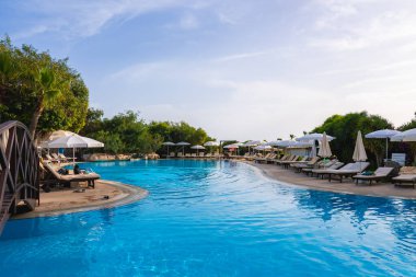 Kıbrıs 'taki bir tatil köyünde berrak mavi su, ahşap bir köprü, güneş pansiyonları, şemsiyeler ve parlak bir gökyüzü altında yemyeşil alanlar yer alıyor..