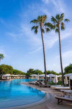 Kıvrımlı kenarlı geniş bir havuz, güneşli şezlonglar, beyaz şemsiyeler ve Kıbrıs 'ta açık mavi bir gökyüzü altında yemyeşil ile çevrili uzun palmiye ağaçları..