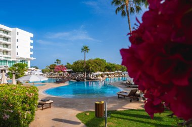 Kıbrıs 'ta bir tatil beldesinde büyük bir havuz, ahşap bir köprü, güneş pansiyonları, pembe çiçekler, palmiye ağaçları ve temiz bir gökyüzünün altında beyaz çok katlı bir bina yer alıyor..