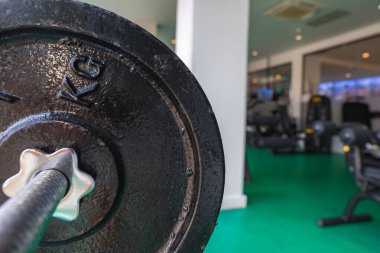 KG işaretli bir ağırlık plakası spor salonundaki bir halterin üzerinde duruyor. Sahne yeşil zemin, egzersiz makineleri ve ışığı yansıtan cam bölme içeriyor..