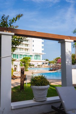 Kıbrıs 'ta yüzme havuzu, güneş pansiyonları, şemsiyeler, beyaz çok katlı bir bina, yeşillik ve temiz bir gökyüzünün altında ahşap bir pergola bulunan modern bir tatil köyü..
