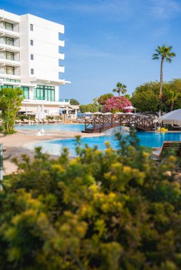 Kıbrıs 'ta bir tatil beldesinde ahşap köprüsü, yemyeşil yeşilliği, pembe çiçekli ağaçları ve berrak mavi gökyüzünün altında modern beyaz bir otel yer alıyor..