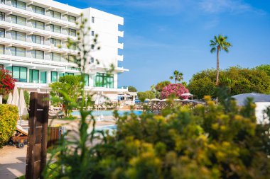 Balkonları, havuzu, yemyeşil otelleri, çiçek açan çalıları, palmiye ağaçları ve Kıbrıs 'ta açık mavi gökyüzü olan modern beyaz çok katlı bir otel..