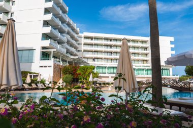 Balkonları, şezlongları, çiçekli bitkileri, ahşap bir köprüsü ve uzun bir palmiye ağacıyla çevrili bir havuzu olan modern beyaz çok katlı bir otel..