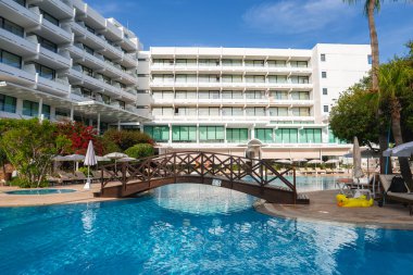 Balkonları olan modern beyaz bir otel ahşap köprüsü olan bir havuza bakıyor. Palmiye ağaçları, güneş pansiyonları, şemsiyeler ve canlı bitkilerle çevrili..