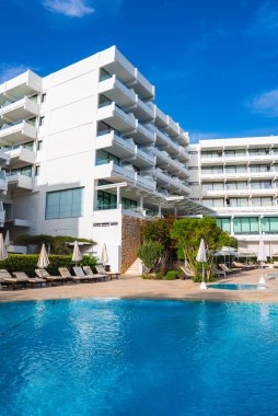 Kıbrıs 'ta balkonları, el değmemiş bir havuzu, şezlongları, şemsiyeleri, yemyeşil ve parlak mavi gökyüzü olan modern beyaz çok katlı bir otel..