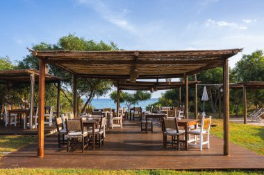 Bir açık hava yemek alanında ahşap pergolalar, kırsal mobilyalar ve arka planda sakin bir deniz manzarası olan yeşillikle çevrili ahşap bir güverte bulunur..