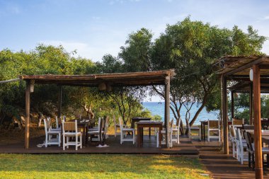 Ahşap masalar, beyaz sandalyeler ve arka planda sakin bir deniz manzaralı yeşillik ile çevrili pergolalar içeren kırsal açık hava yemek alanı..