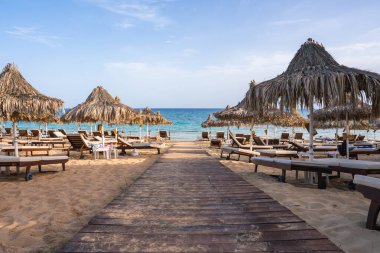 Kıbrıs 'taki kumlu bir plajda açık mavi gökyüzünün altında ahşap bir patika, turkuaz sular, sıralanmış güneş şezlongları ve sazdan şemsiyeler bulunuyor..
