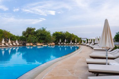 Berrak mavi sularla çevrili geniş kıvrımlı bir havuz. Beyaz güneş şezlongları ve şemsiyeler, taşlarla döşenmiş havuz kenarında, sakin bir ortamda..