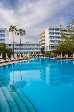 Kıbrıs 'ta büyük bir yüzme havuzu, beyaz binalar, balkonlar, palmiye ağaçları, yemyeşil ve dağınık bulutlu açık mavi bir gökyüzü olan modern bir tatil köyü..