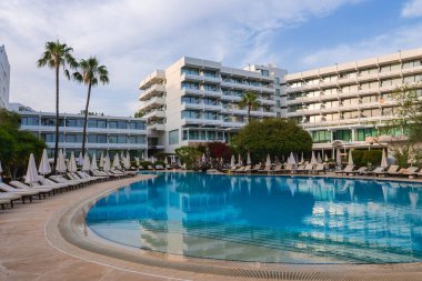 Kıbrıs 'ta geniş kavisli havuzu, güneşli odaları, şemsiyeleri, yüksek palmiyeleri ve açık gökyüzü altında beyaz çok katlı binaları olan modern bir otel..