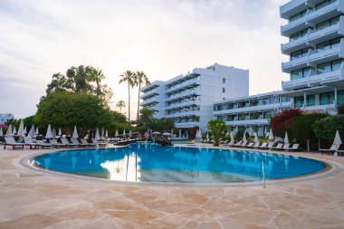 Büyük yuvarlak bir havuz, şezlonglar, şemsiyeler ve dekoratif bir köprü, palmiye ağaçları ile çevrili ve modern beyaz otel binaları tarafından gözden kaçırılmış..