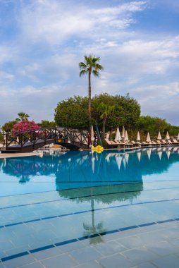Kıbrıs tatil beldesinde sakin bir havuzda ahşap bir köprü, uzun bir palmiye ağacı, beyaz şemsiyeli güneşli şezlonglar, yemyeşil ve pembe begonviller yer alıyor..