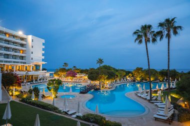 Kıbrıs 'ta ışıklı bir yüzme havuzu, şezlonglar, palmiye ağaçları, bougainvillea, modern bir otel ve arka planda deniz bulunan bir tatil beldesinin akşam manzarası.