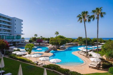 Kıbrıs 'ta bir tatil beldesinde güneş pansiyonları, beyaz şemsiyeler, modern bir otel, palmiye ağaçları ve Akdeniz' e bir bakış bulunan bir kıvrımlı havuz yer alıyor..