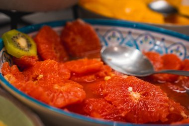 Kırmızı turuncu turunçgil dilimleri ve kiviyle dekoratif bir kase, desenli bir kenarı var. Metal bir kaşık içinde duruyor, arka planda bulanık yiyecek parçaları var..