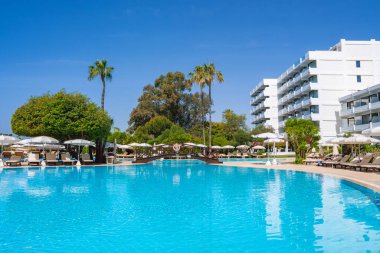 Kıbrıs 'ta büyük bir havuzu, dinlenme yerleri, şemsiyeleri, beyaz çok katlı bir otel, palmiye ağaçları, ahşap bir köprü ve parlak mavi bir gökyüzü olan bir tatil köyü..